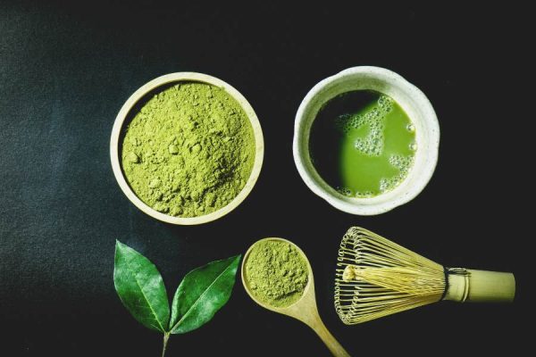 A cup of matcha beside a bowl of powder, spoon, and whisk